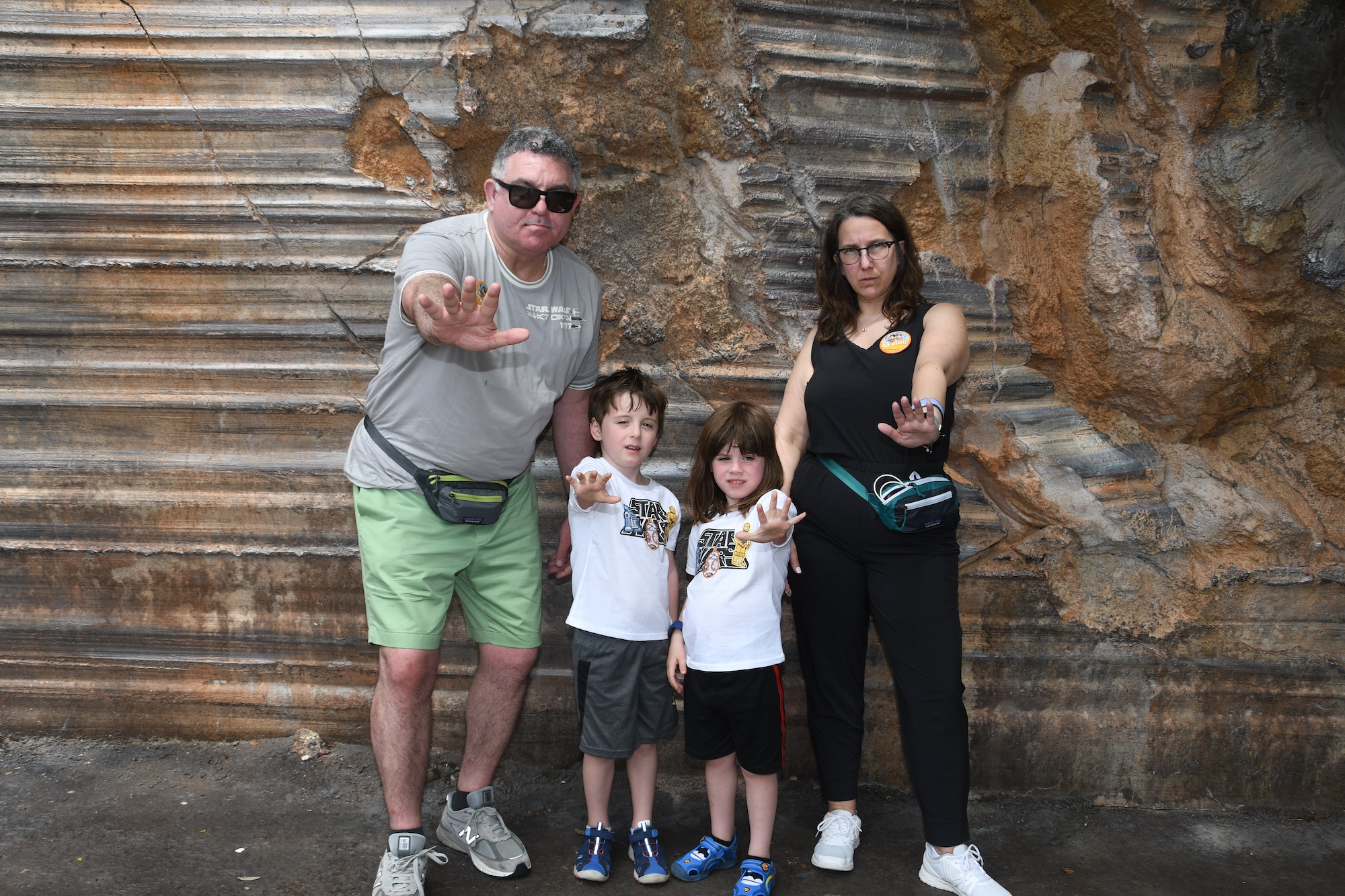 A man, woman, and two children strike serious poses in front of a rock wall. The adults sport stylish sunglasses and fanny packs while the children wear matching white shirts and blue shoes. They look like the perfect clients for a luxury family travel agent.